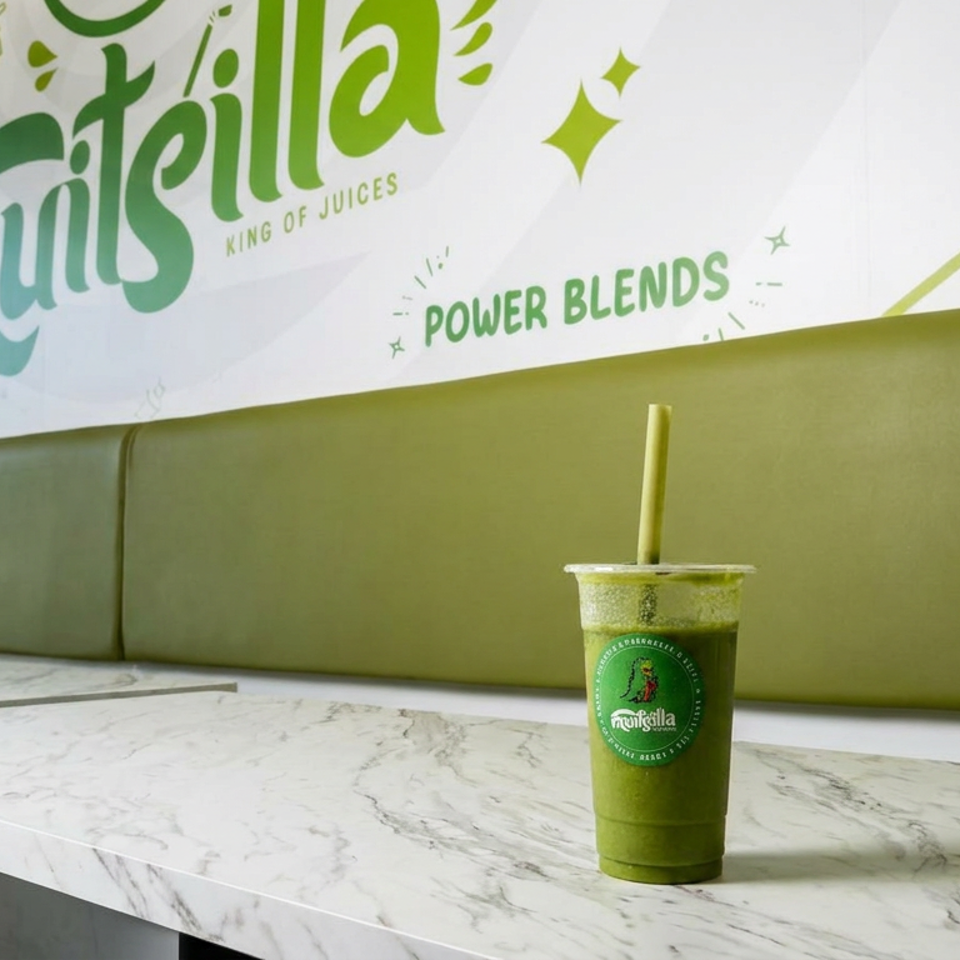 Green smoothie with a straw on a marble table inside a Fruiteilla juice shop, beneath a colorful Power Blends wall graphic.
